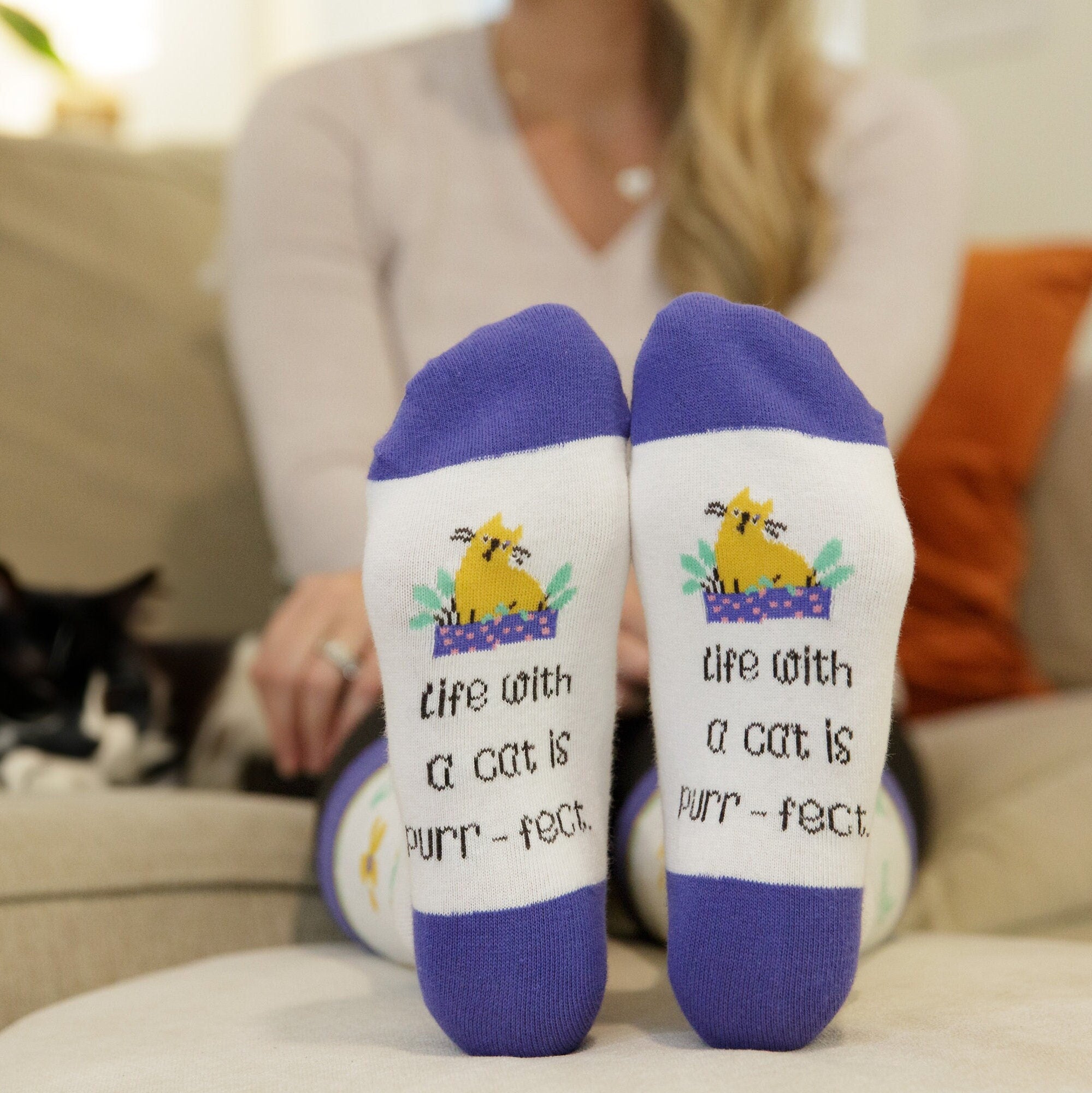 Person wearing socks with a cat-themed design and text, sitting on a couch with a cat in the background.